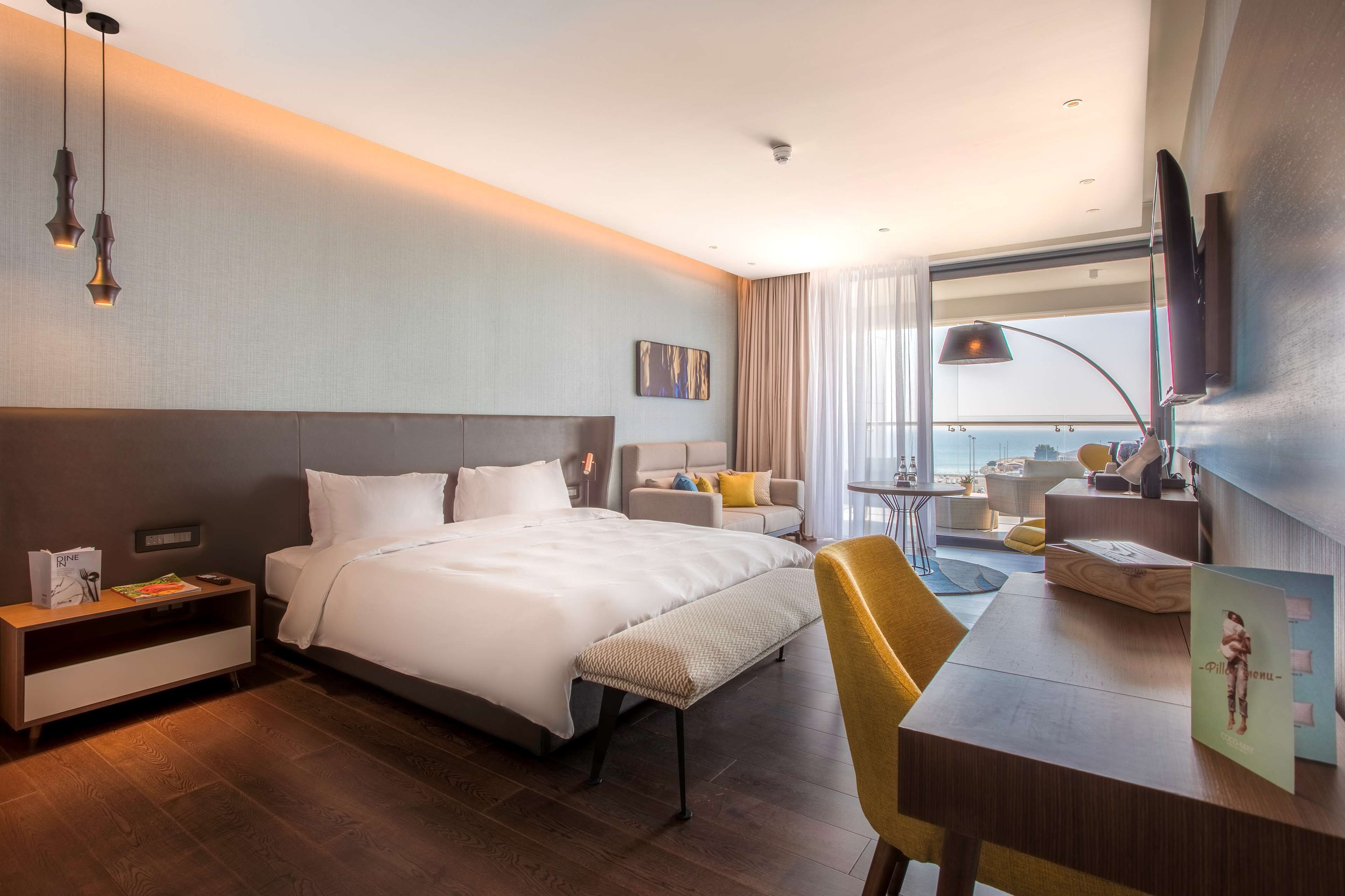 A modern hotel room with a large bed, bedside table, and a desk with a chair. There is a TV mounted on the wall and a window with curtains.