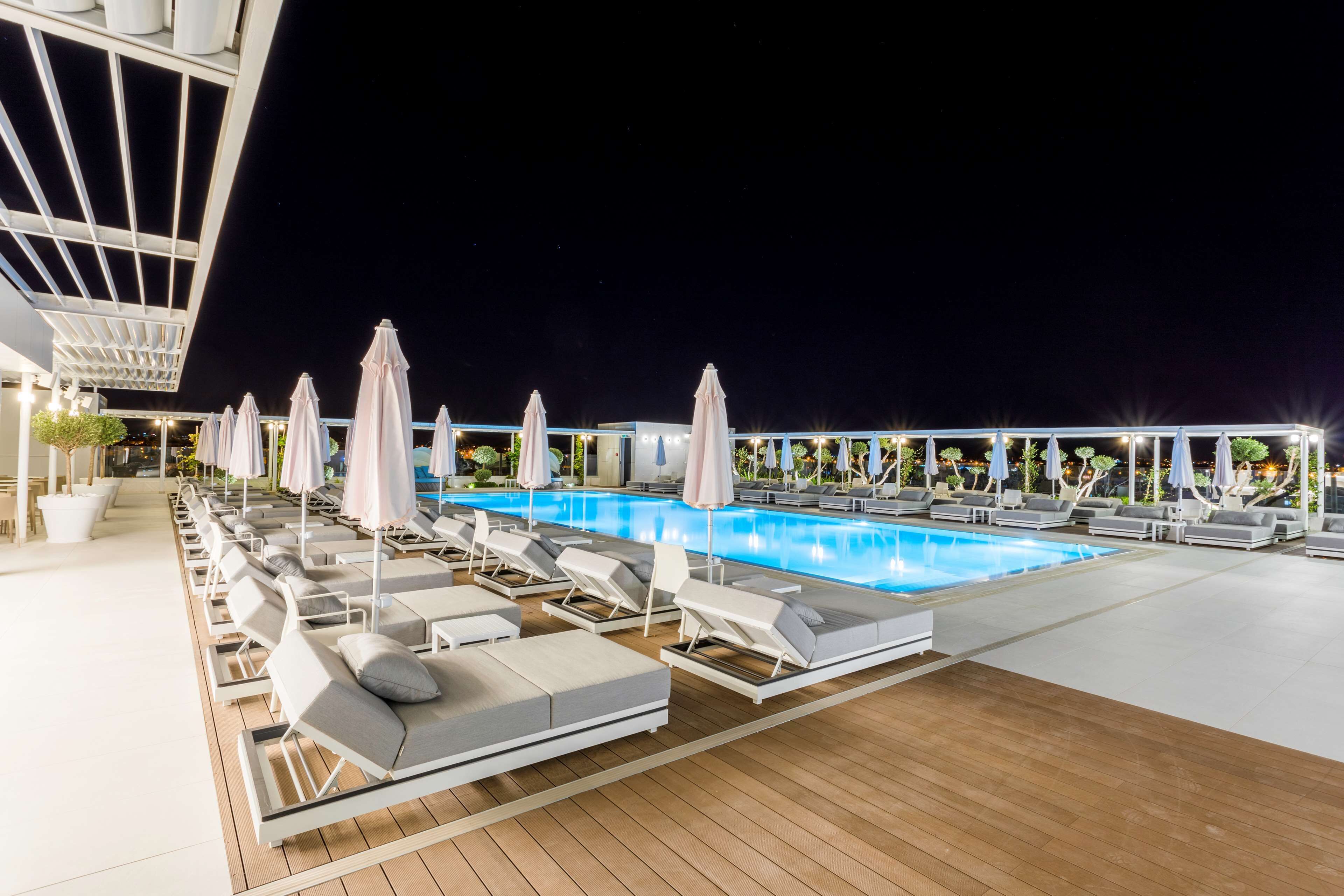 Un espace aménagé au bord de la piscine, avec des chaises longues et des parasols, une terrasse en bois et une piscine à l’eau d’un bleu limpide.
