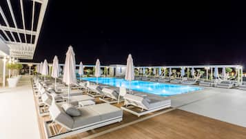 Un espace aménagé au bord de la piscine, avec des chaises longues et des parasols, une terrasse en bois et une piscine à l’eau d’un bleu limpide.