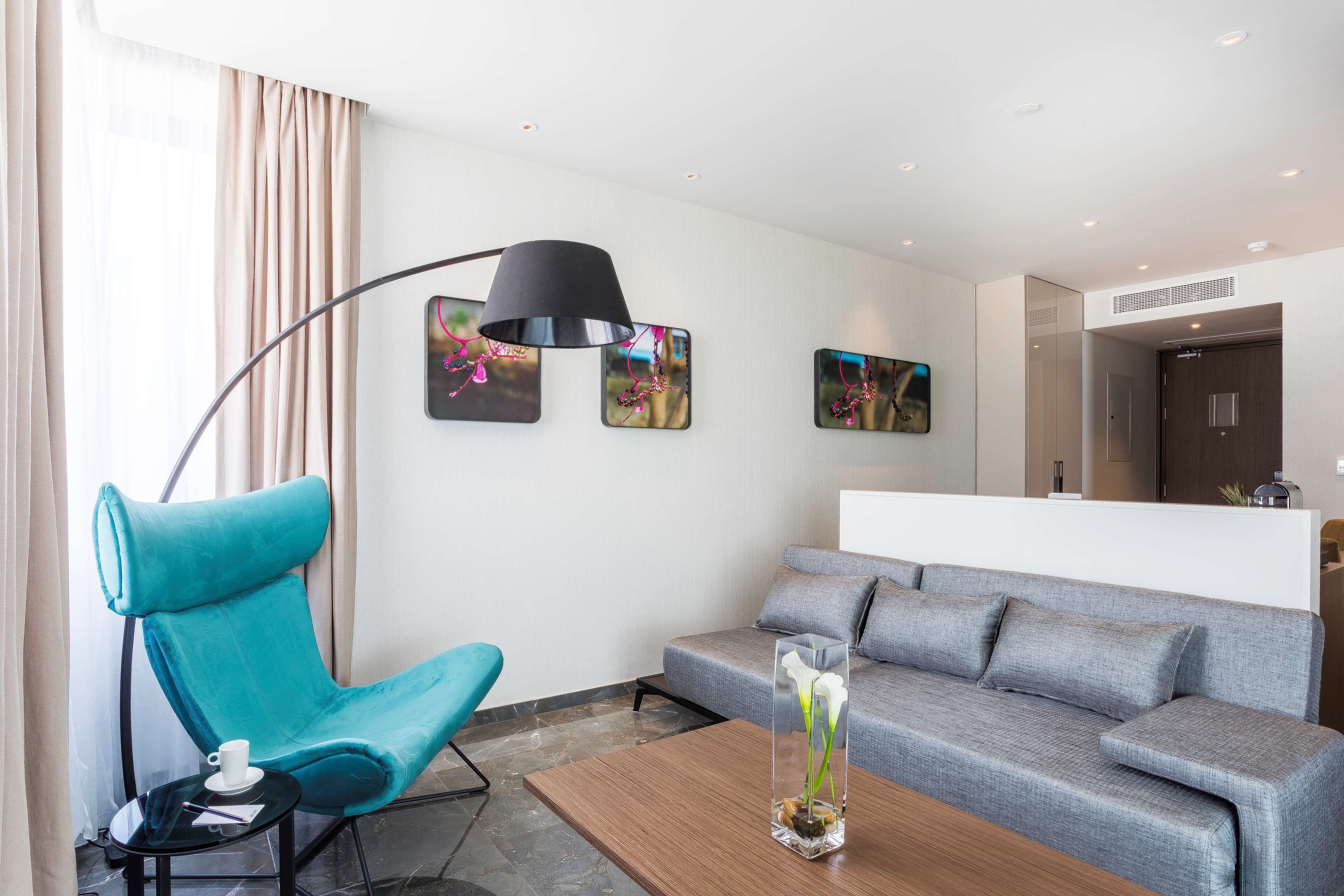 Une chambre d’hôtel moderne avec un fauteuil turquoise, un canapé gris, une table basse agrémentée d’un vase et un lampadaire noir.