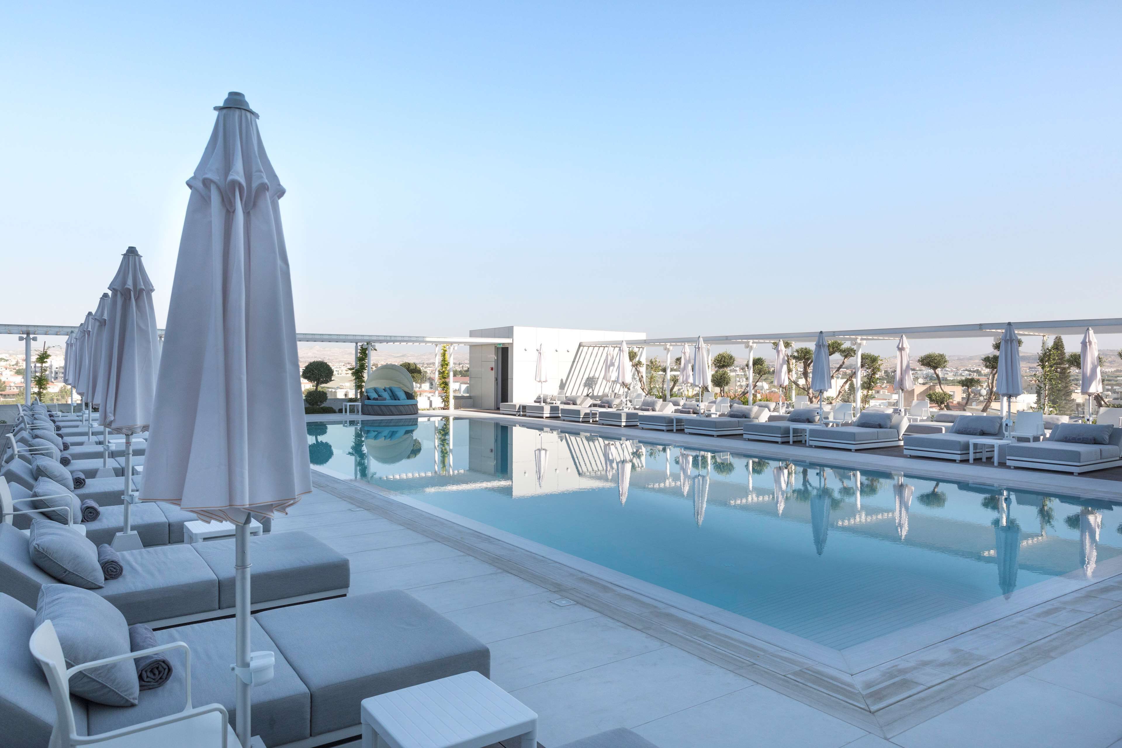 A rooftop pool area with lounge chairs and umbrellas, a clear sky, and a view of the cityscape.