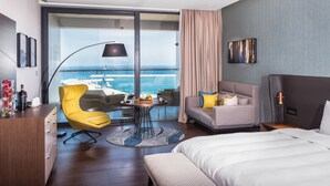 A modern hotel room with a large bed, a sofa, a dining table with fruit, a yellow chair, and a view of the ocean.
