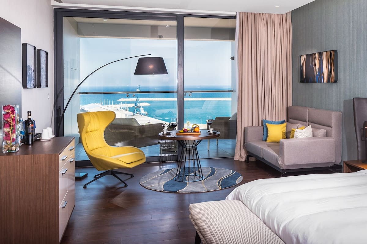 A modern hotel room with a large bed, a sofa, a dining table with fruit, a yellow chair, and a view of the ocean.
