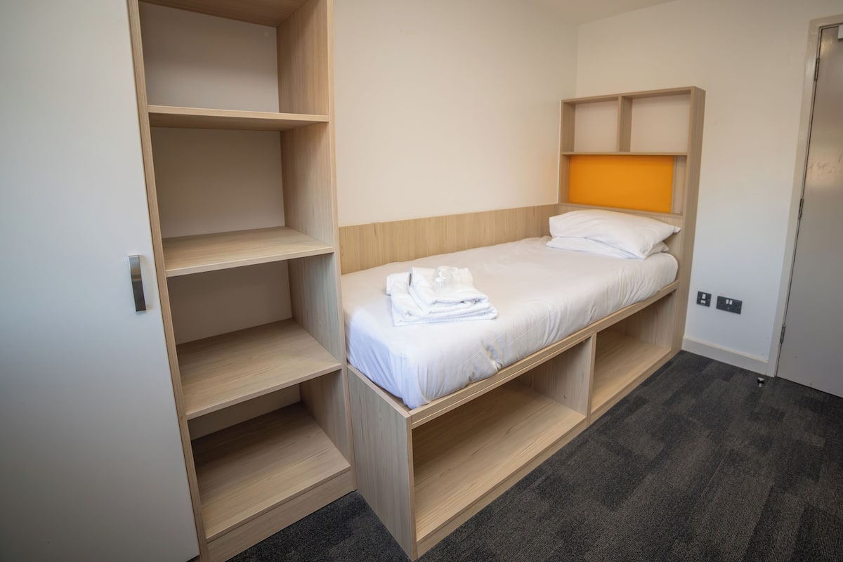 A compact room with a single bed, built-in shelving, and a wardrobe.
