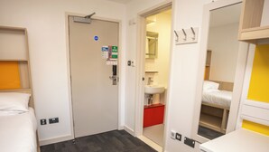 A hotel room with a bed, a desk, a chair, a wardrobe, a mirror, a sink, and a door leading to another room.