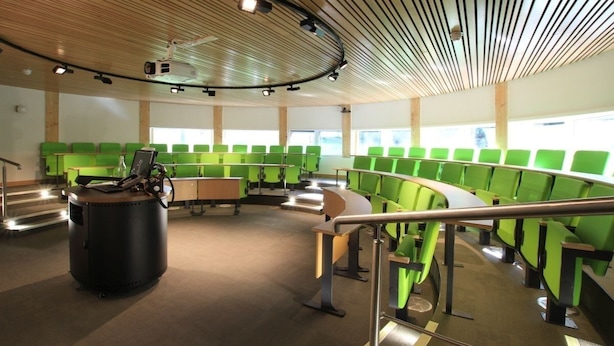 A modern conference room with green seating arranged in a semi-circle, a podium, and a projector mounted on the ceiling.