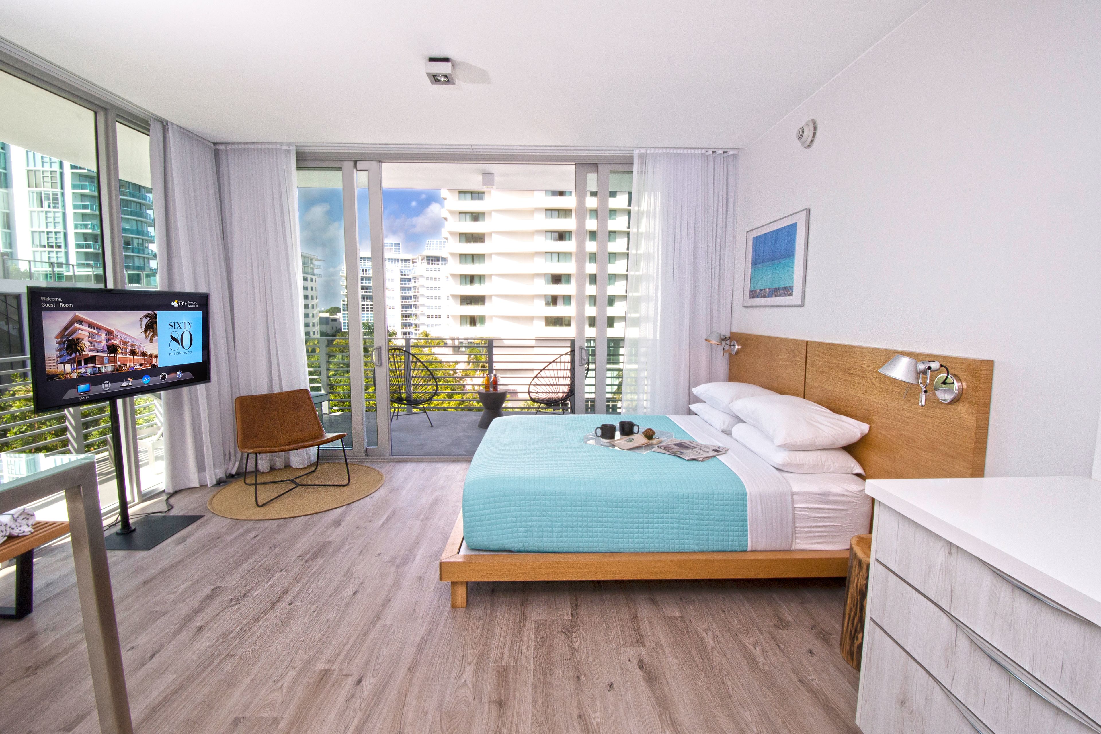A modern hotel room with a large bed, a TV, a balcony with a view of buildings, and a wooden headboard.