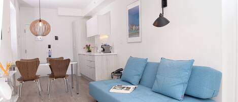 Ein modernes Wohnzimmer mit einem blauen Sofa, zwei Holzstühlen, einem Glastisch und einer Küche im Hintergrund.