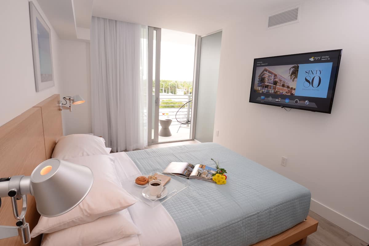 Una habitación de hotel con una cama, una mesita de noche con lámpara, una ventana con cortinas y un televisor mostrando una imagen del hotel.