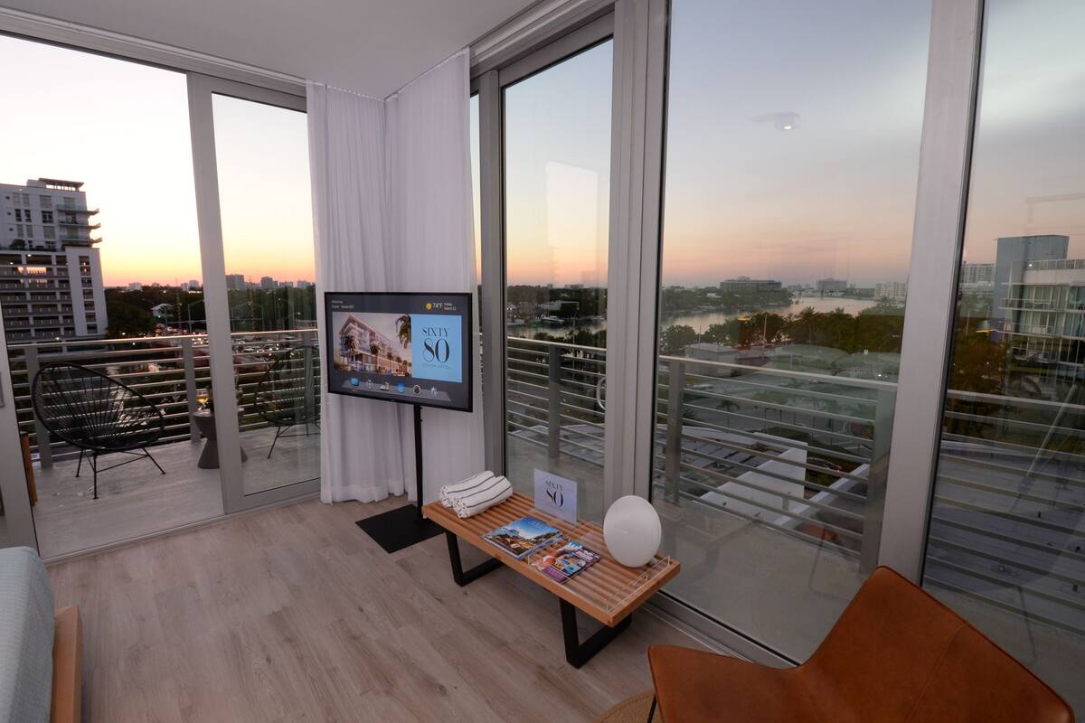 Una sala de estar moderna con un televisor grande, una mesa de centro de madera con revistas, un sillón cómodo y vistas de la ciudad a través de amplios ventanales.