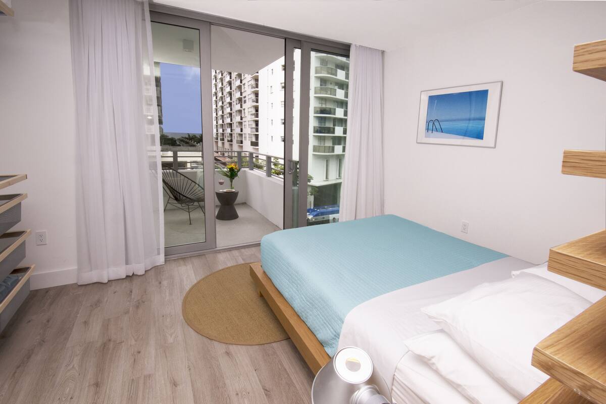 Una habitación de hotel moderna con una cama, una mesita, una silla, vistas al balcón y un cuadro en la pared.