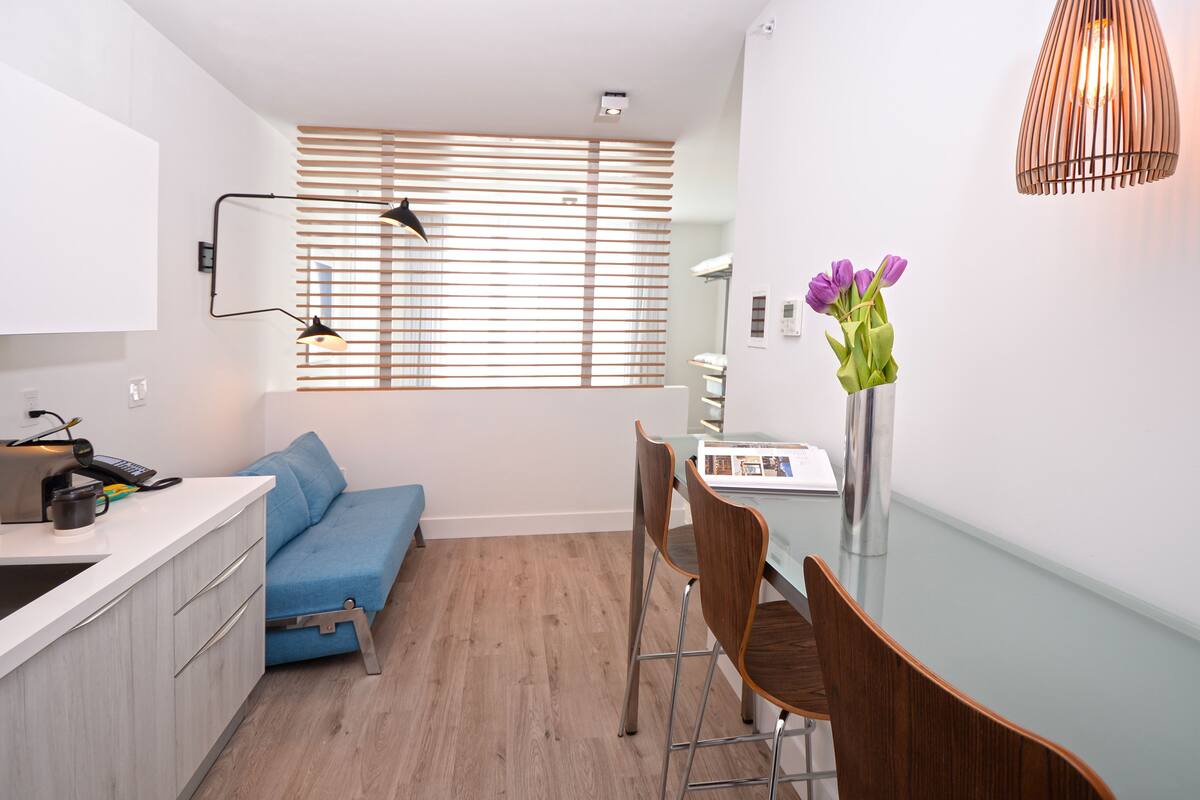 Una cocina compacta con un sofá azul, sillas de comedor de madera y un jarrón con flores moradas.
