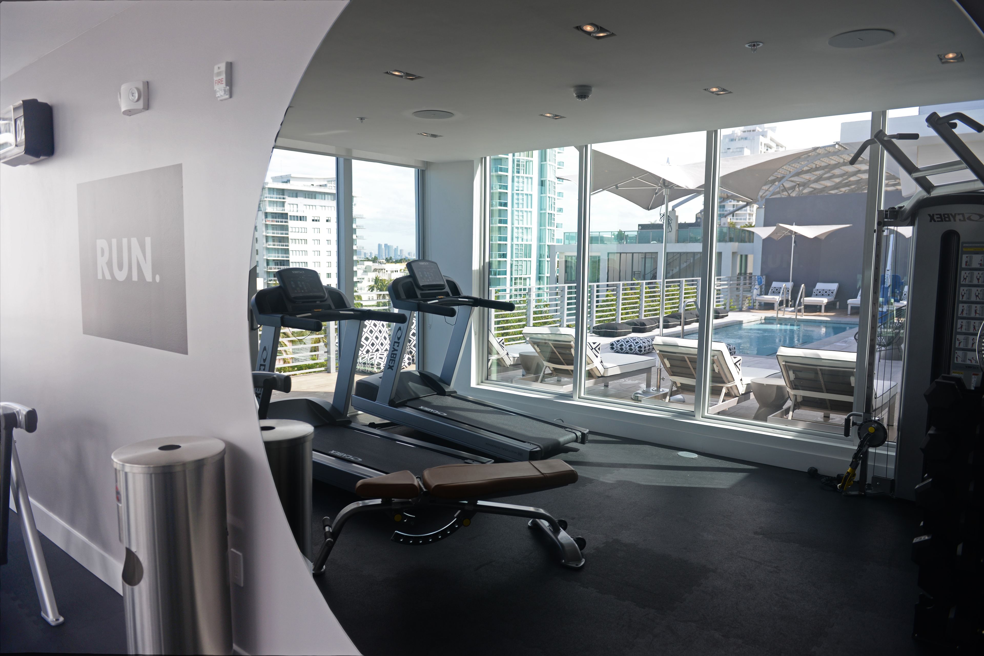 Ett gym med löpband, en bänk och utsikt över en pool och stadssilhuetten genom stora fönster.