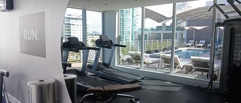 Ett gym med löpband, en bänk och utsikt över en pool och stadssilhuetten genom stora fönster.