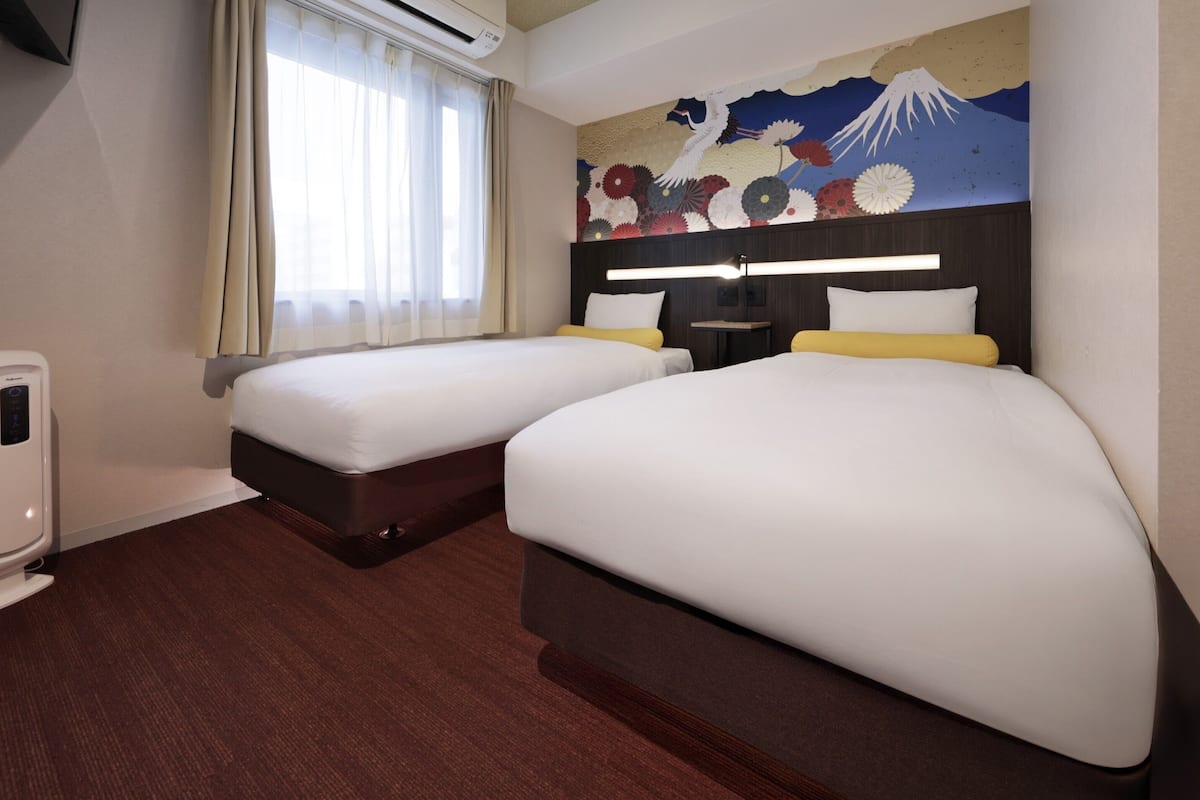 A hotel room with two beds, a wall-mounted air conditioner, a bedside table with a lamp, and a large painting of Mount Fuji.
