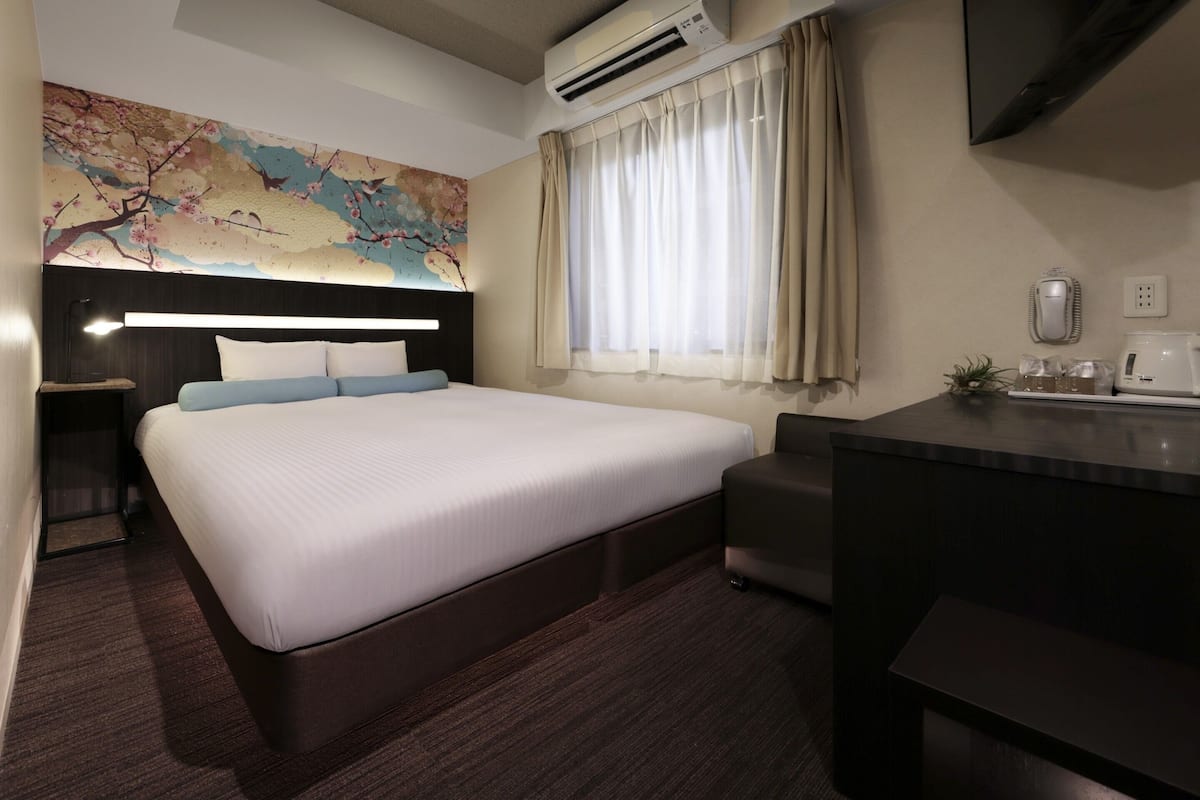 A hotel room with a bed, bedside table, desk, air conditioning unit, and a large window with curtains.