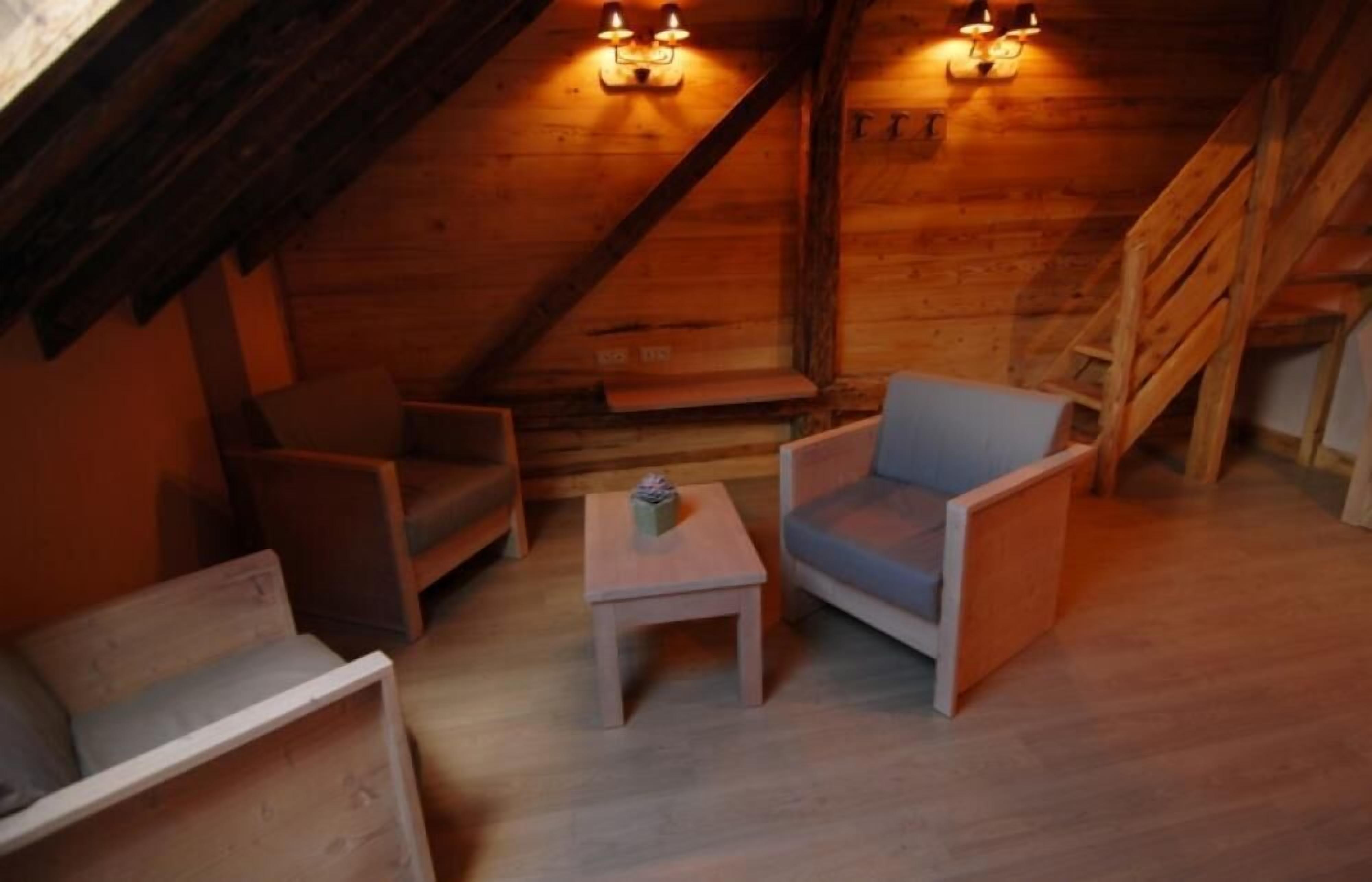 A cozy attic room with wooden walls and a sloped ceiling, featuring two armchairs, a small table, and a staircase.