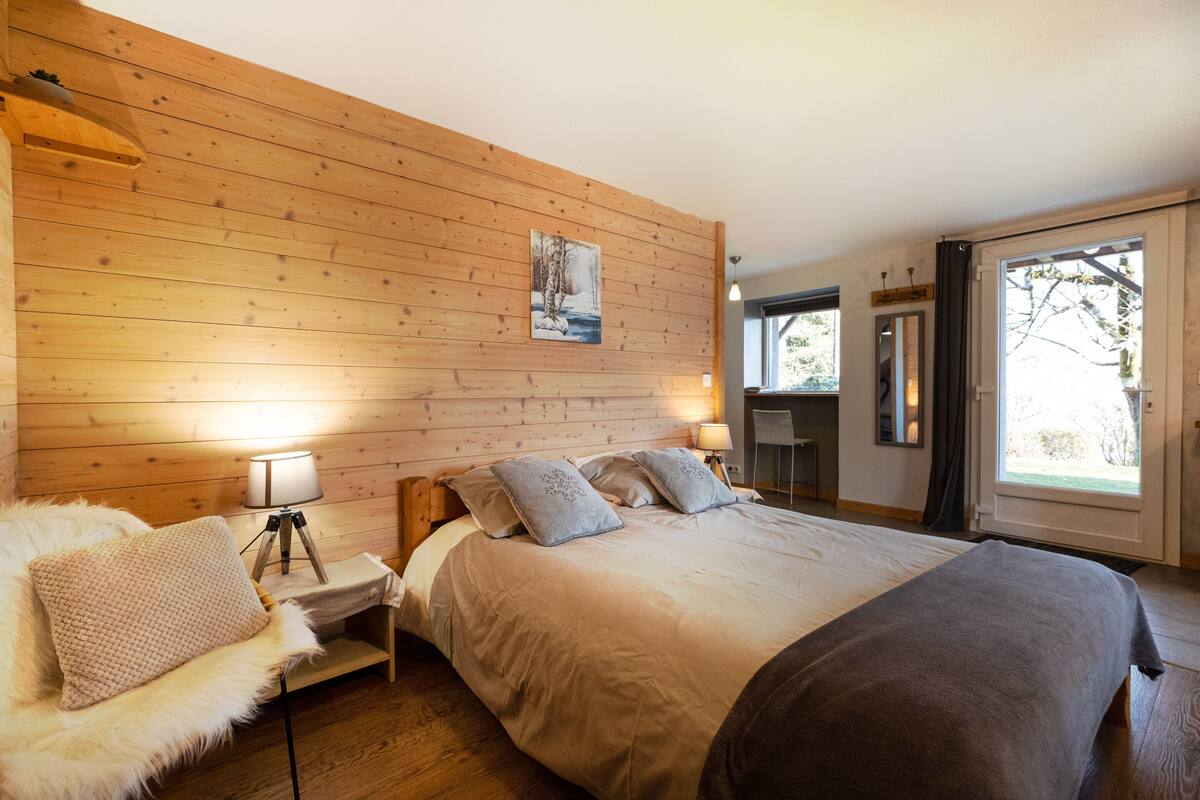 Une chambre confortable avec une tête de lit en bois, un grand lit recouvert d’une couverture grise, un fauteuil moelleux et une lampe de chevet.