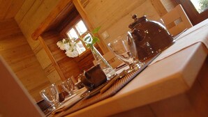 Un intérieur en bois avec une table à manger dressée pour deux personnes, agrémentée d’une carafe en verre transparent, d’un vase avec des fleurs blanches et d’une chaise en bois.