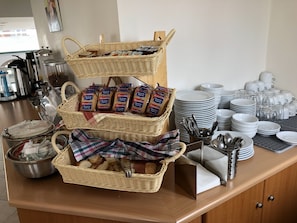 A buffet setup with wicker baskets, bread, and stacked white plates and cups. - Ageri (Tinos)
