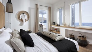 A modern bedroom with a large bed, a hanging light fixture, a window with a view of the sea, and a small table with a basket. - Villa Delmar - Delmar Collection (Milos)