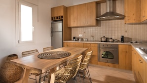 A modern kitchen with wooden cabinets, a stainless steel oven, and a dining area with a wooden table and wicker chairs. - Villa Delmar - Delmar Collection (Milos)
