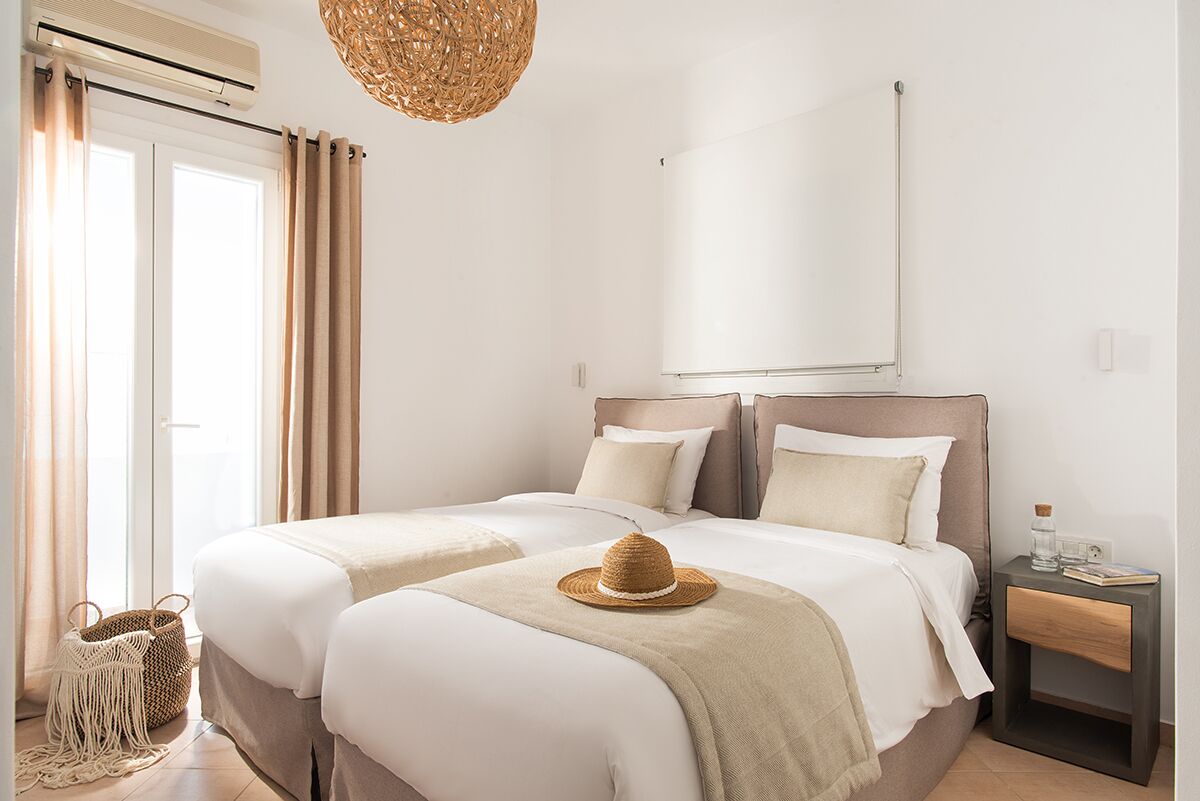 A hotel room with two beds, a large window with curtains, a woven light fixture, and a bedside table with a book and a bottle.
