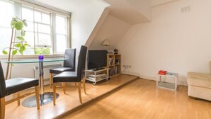 Uma sala de estar moderna com uma mesa de vidro, cadeiras pretas, uma televisão, uma estante e um pequeno sofá.