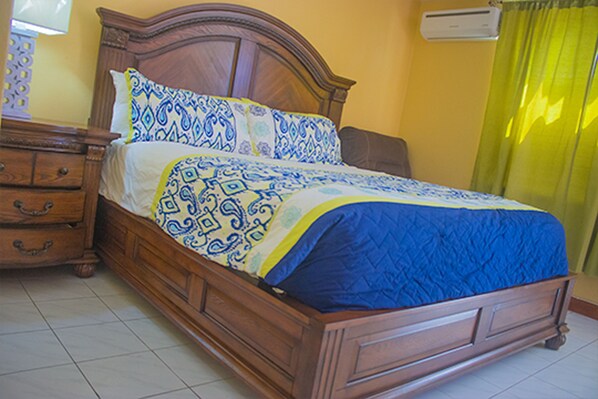 A wooden bed with patterned bedding, a nightstand, and a wall-mounted air conditioner.