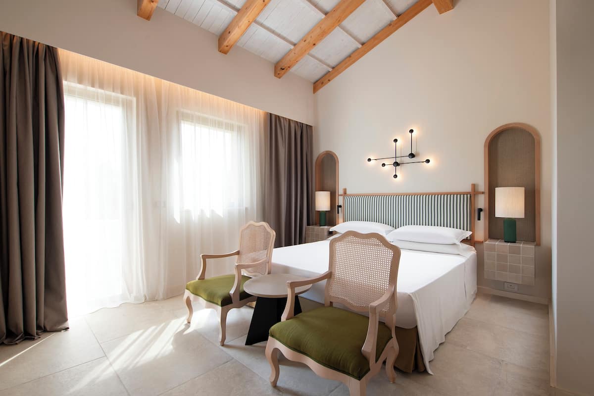 A hotel room with a bed, two chairs, a small table, a bedside lamp, a wall-mounted light fixture, and a window with curtains.