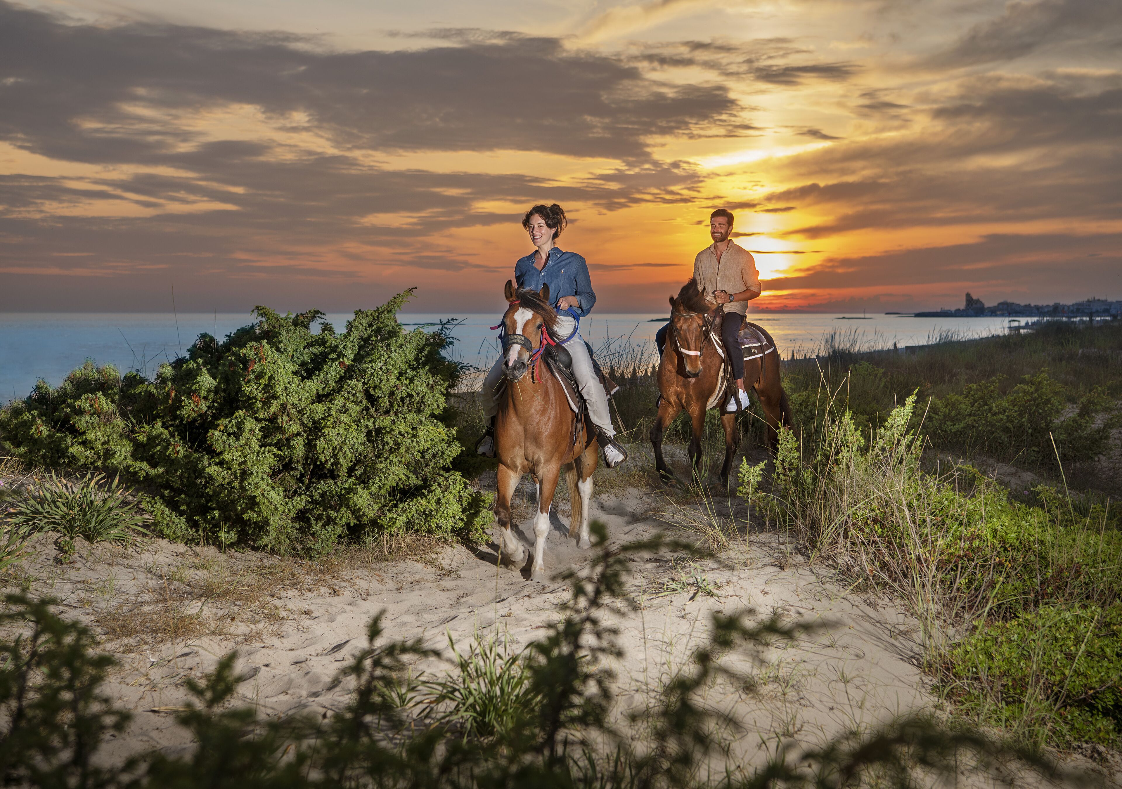 Two people riding horses on a sandy path with vegetation on either side, overlooking a body of water at sunset.