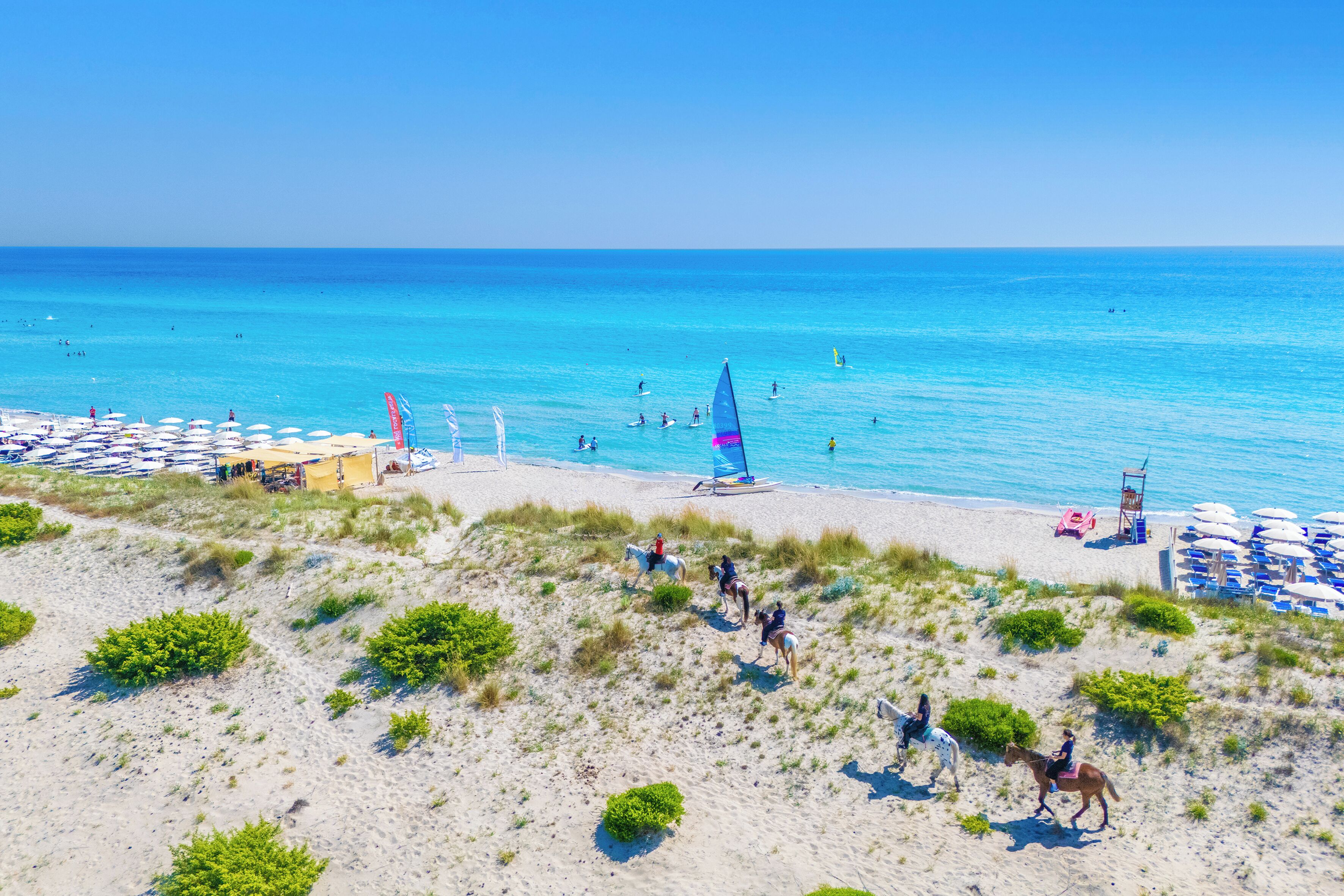 A beach with a sandy area, a sailboat, and people on horses.