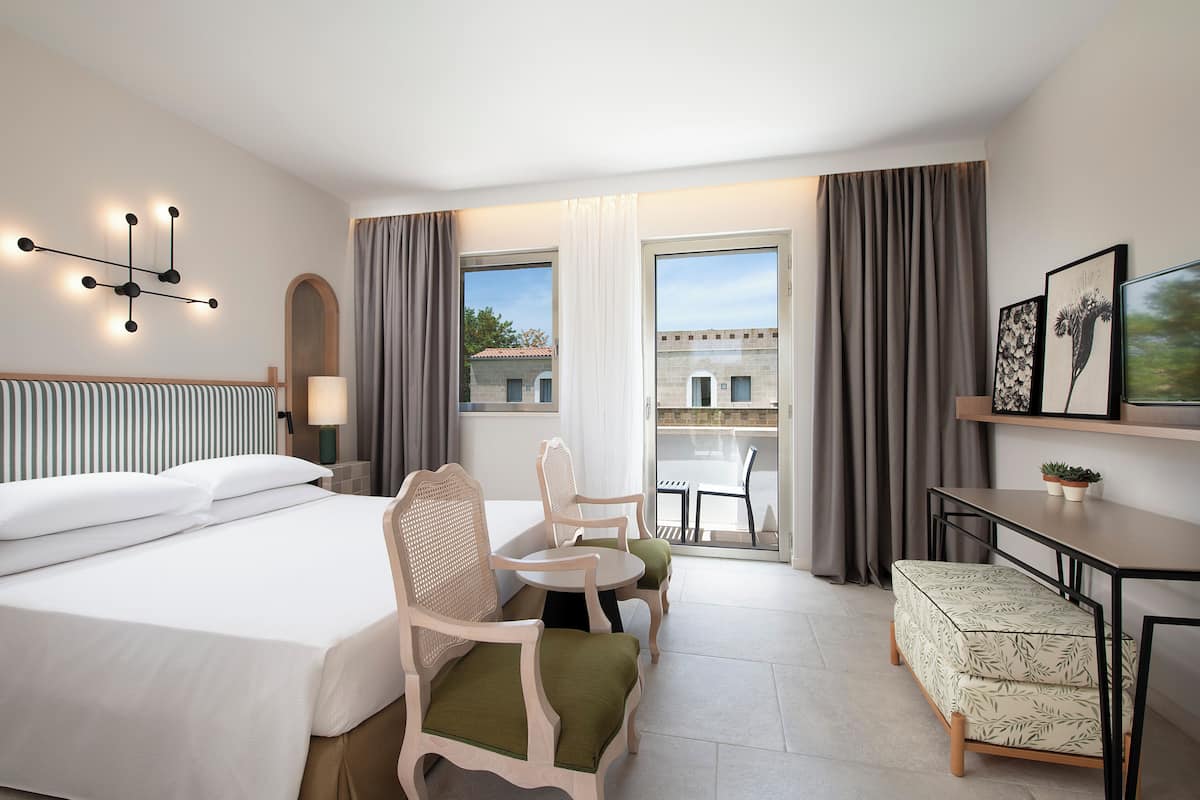 A hotel room with a bed, two chairs, a small table, a bench, a desk, and a view through the window.