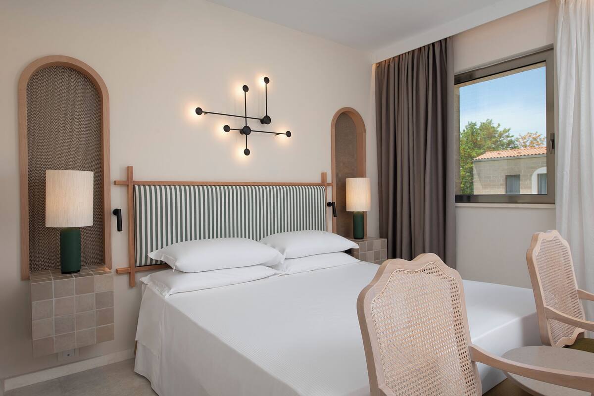 A hotel room with a bed, two bedside tables with lamps, a chair, a wall-mounted light fixture, and a window with curtains.
