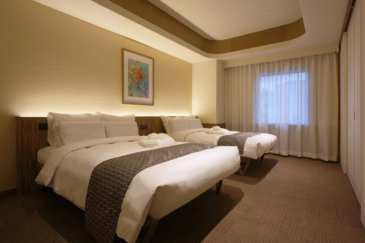 A hotel room with two beds, a painting on the wall, and a large window with curtains.