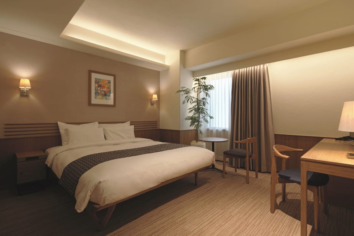 A hotel room with a bed, bedside lamps, a desk, a chair, a small table, and a potted plant.