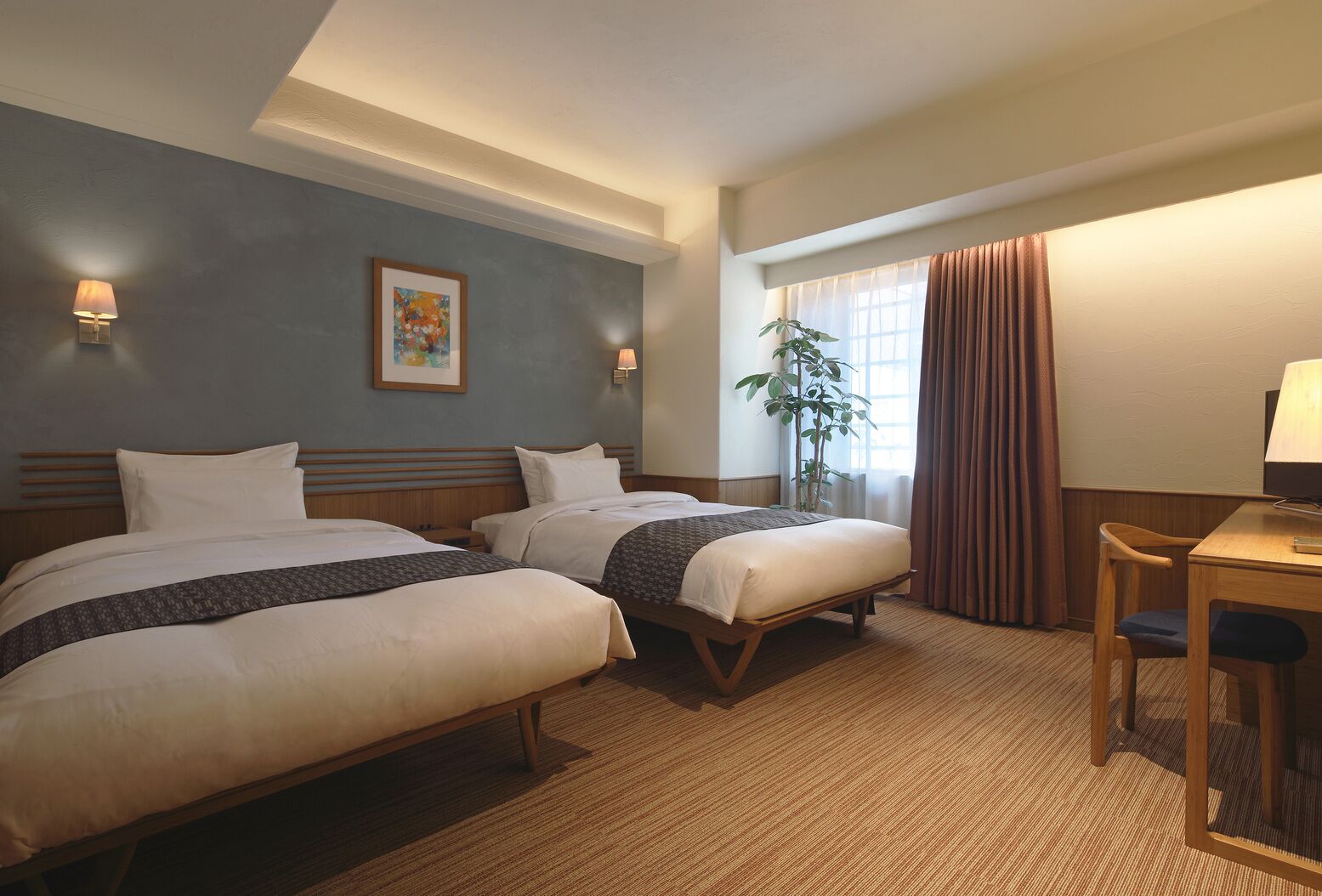 Habitación de hotel con dos camas, un escritorio, una silla, una lámpara, una planta y un cuadro en la pared.