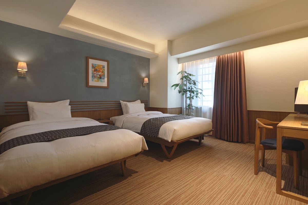 A hotel room with two beds, a desk, a chair, a lamp, a plant, and a painting on the wall.