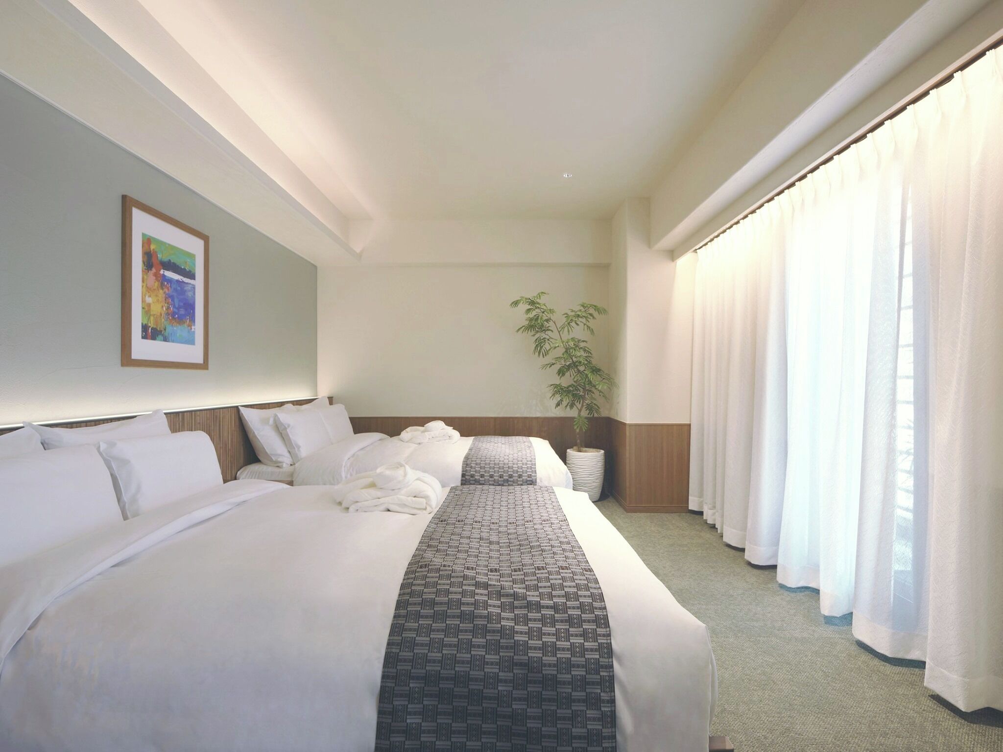 A hotel room with a large bed, a bedside table, a potted plant, and a window with curtains.