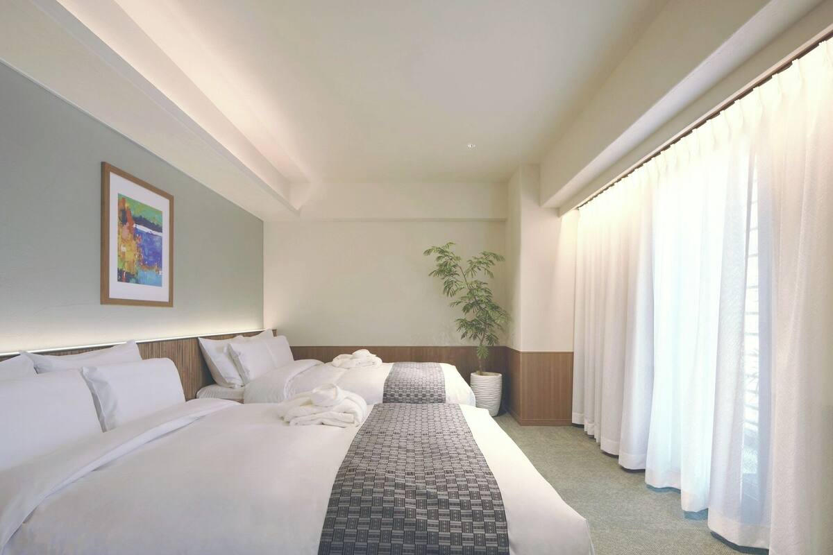 A hotel room with a large bed, a bedside table, a potted plant, and a window with curtains.