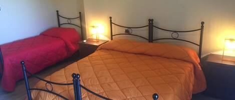 A double bed with a metal frame and a quilted orange bedspread. There are two bedside tables with lamps, and a red bedspread is visible on the left.