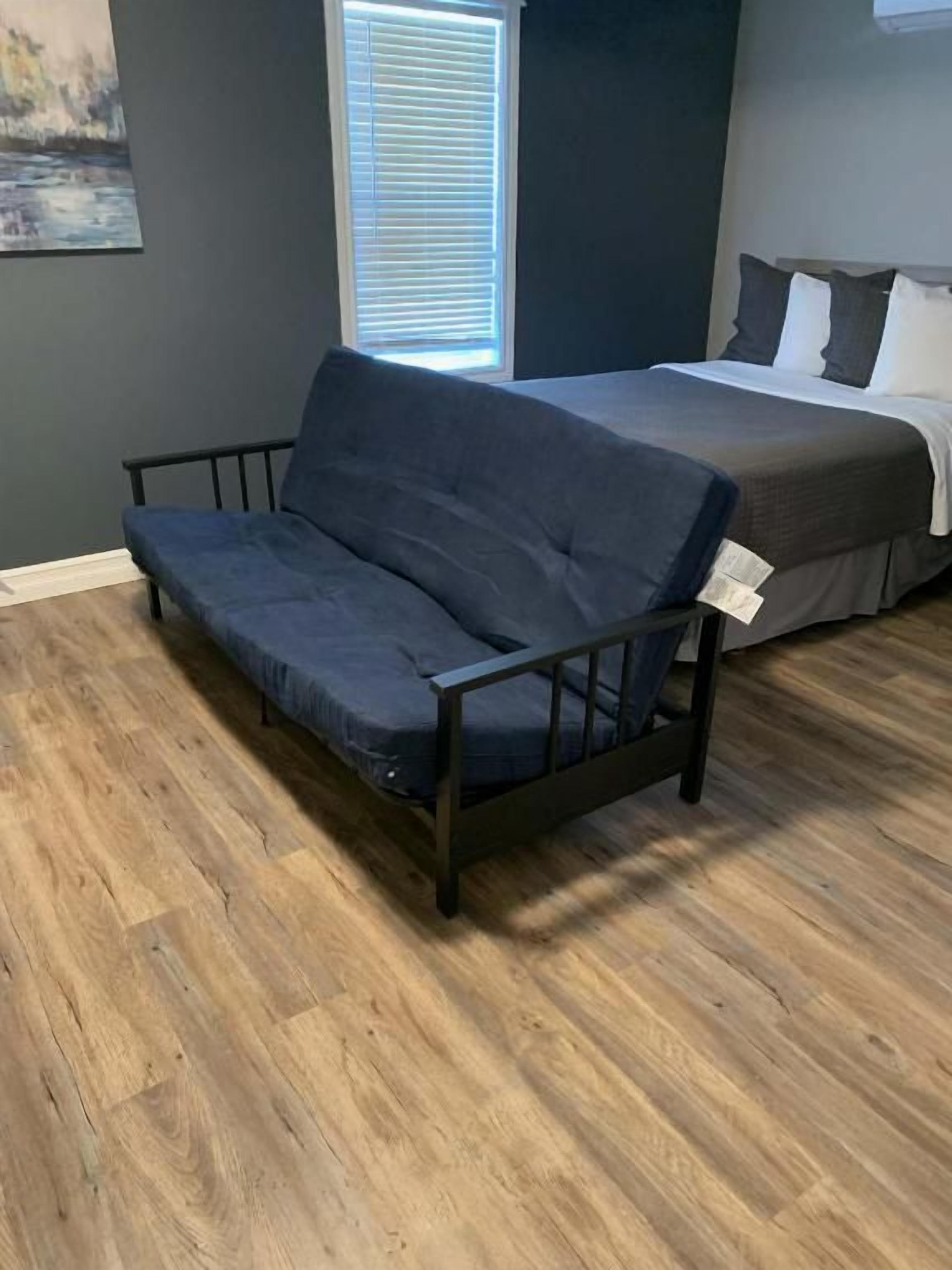 A room with a dark blue sofa, a bed with white bedding, and a window with blinds.
