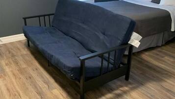 A room with a dark blue sofa, a bed with white bedding, and a window with blinds.