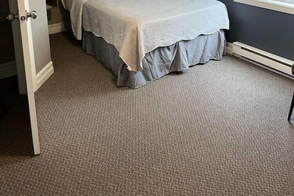 Un dormitorio con cama, unidad de aire acondicionado, ventana con persianas y suelo alfombrado.
