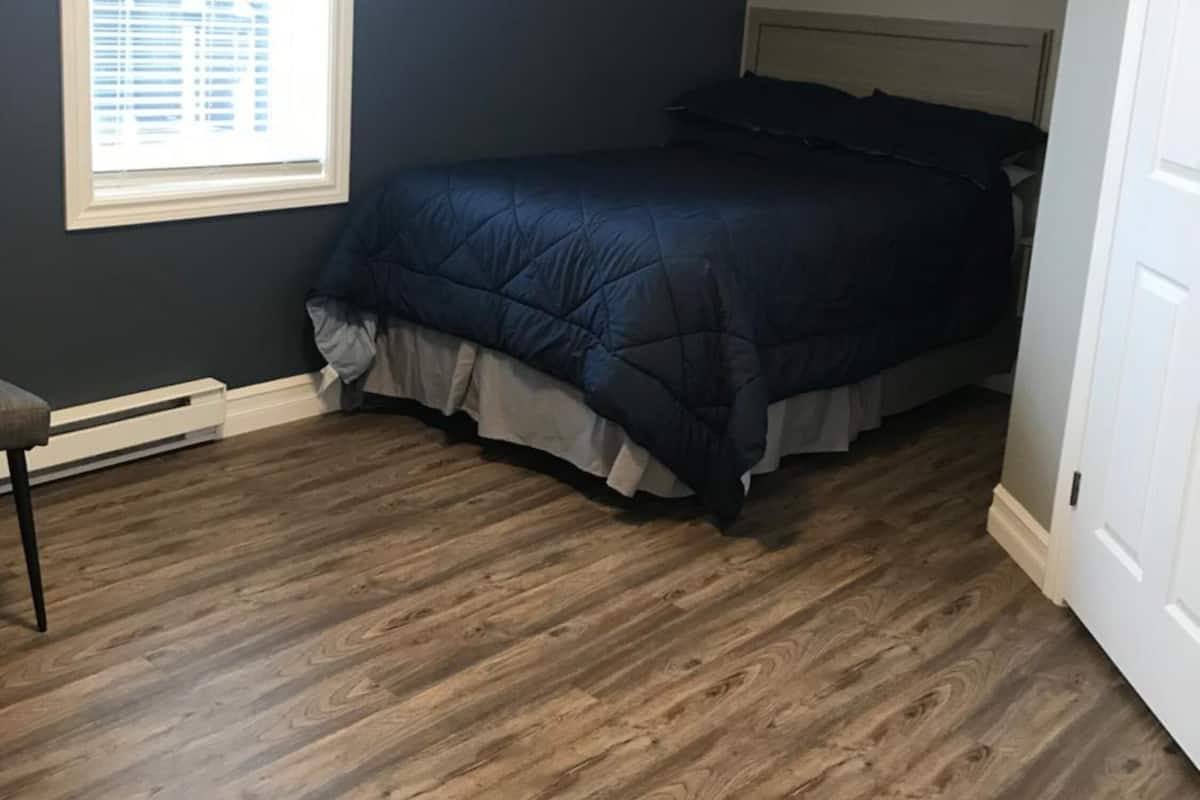 Un dormitorio con pared azul oscuro, suelo de madera, cama con colcha azul oscuro, ventana con persianas y un equipo de aire acondicionado.