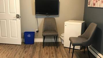 Una habitación con un televisor montado en la pared, un electrodoméstico blanco pequeño, una silla gris, un bote azul para reciclaje y una puerta con el número 3.