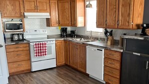 A modern kitchen with wooden cabinets, a white stove, a dishwasher, and a black garbage bin. - Stanley House (Clarenville)