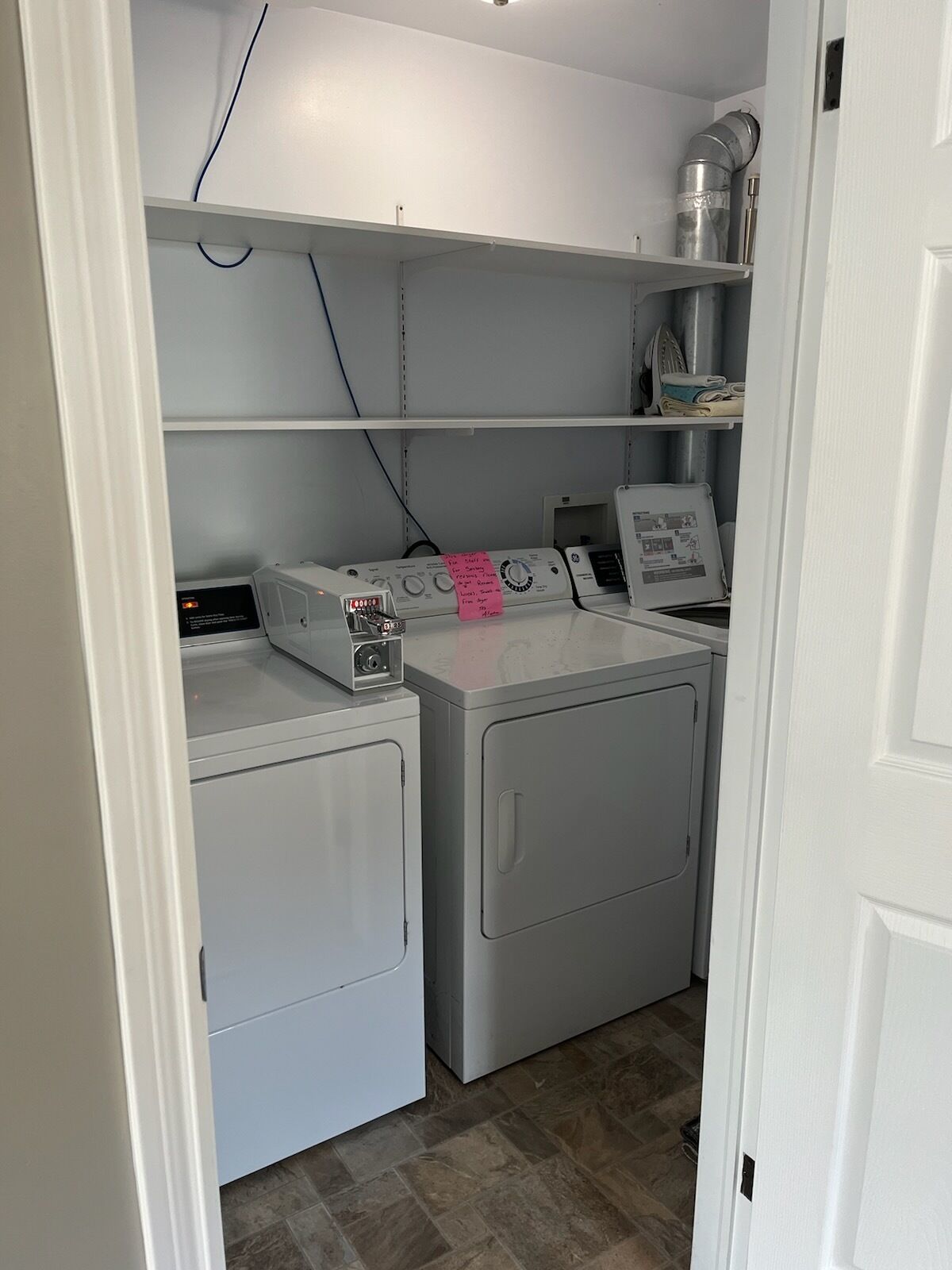 A laundry room with two front-loading washing machines, a dryer, and a shelf above containing a water heater and other items.