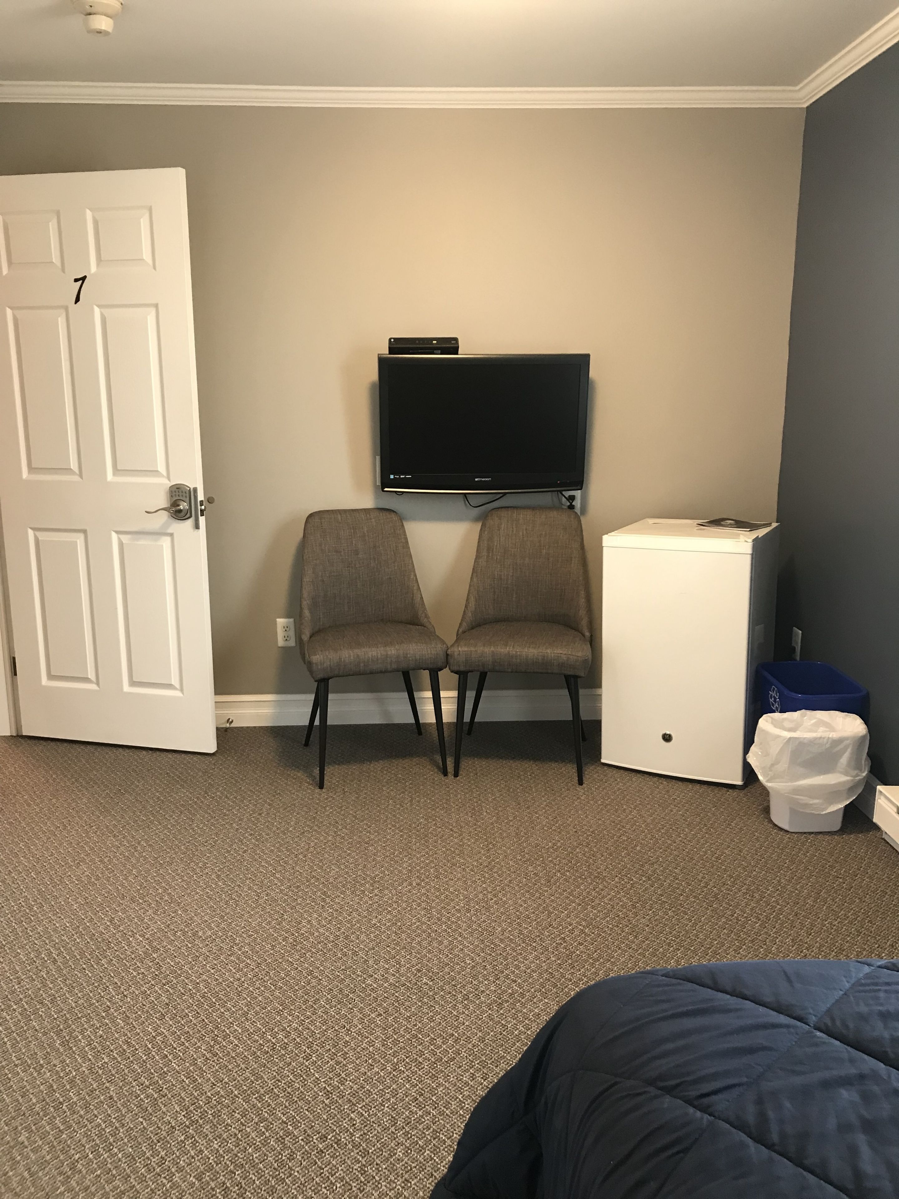 A room with a door, two chairs, a small white cabinet, a television, and a trash bin.