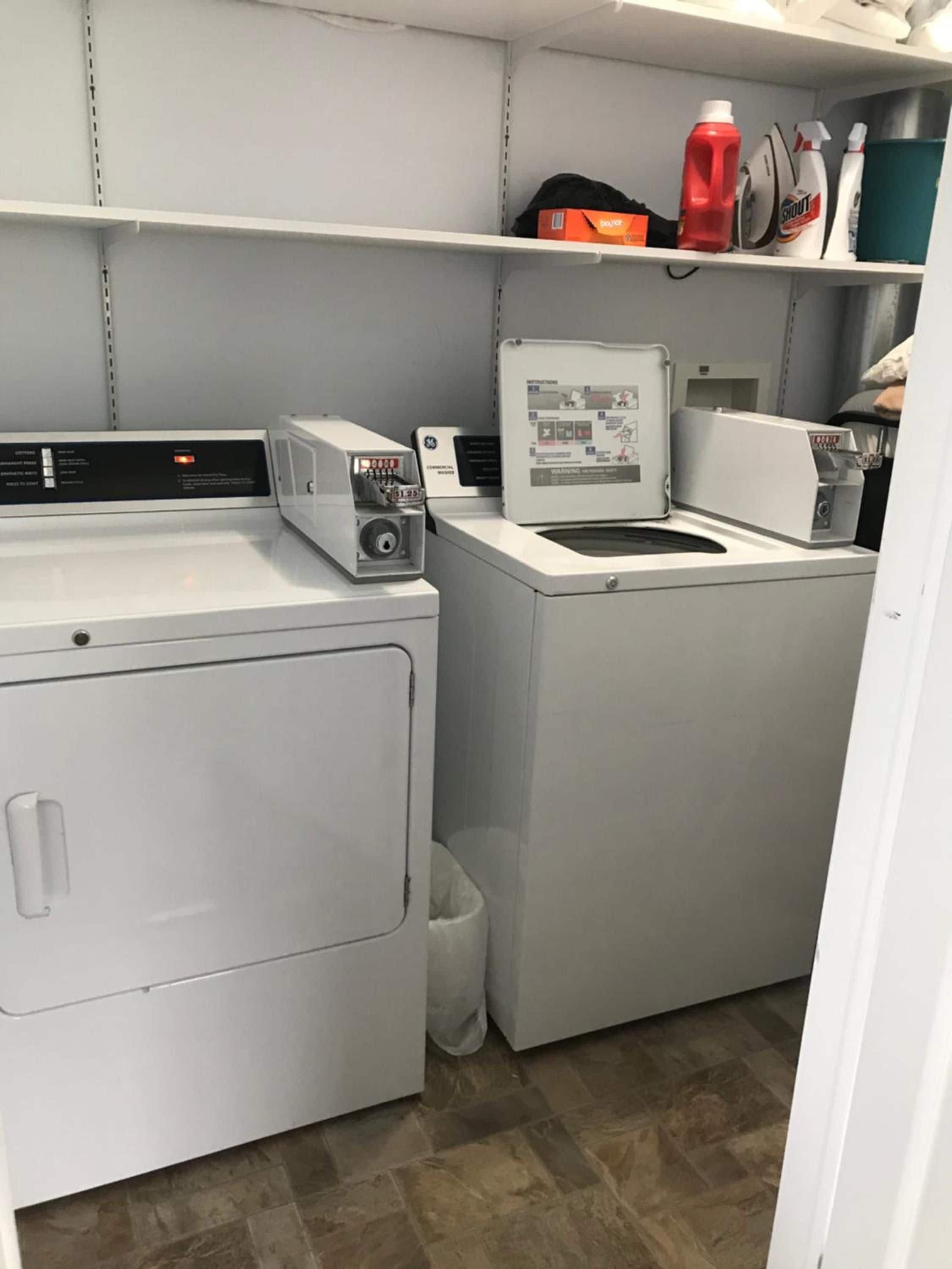 A laundry room with two white washing machines, a dryer, and a shelf with cleaning supplies.
